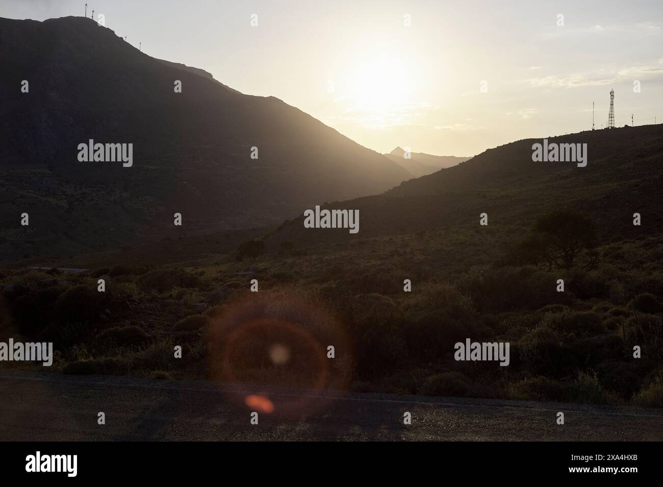 Ein ruhiger Sonnenuntergang hinter den Bergen, mit einem warmen Licht und einer Linse, eine Straße im Vordergrund, die teilweise vom Sonnenlicht beleuchtet wird. Stockfoto