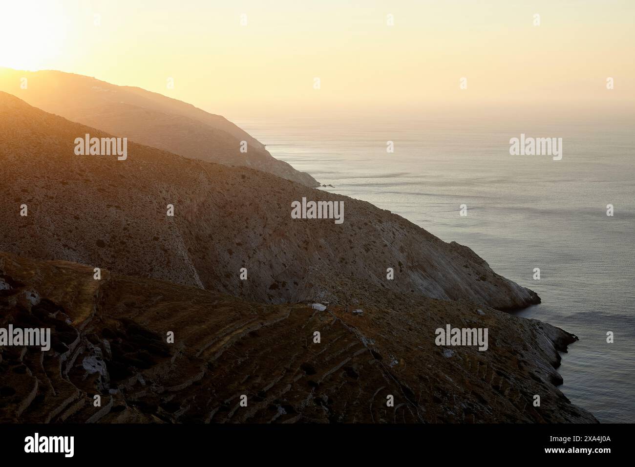 Das Bild zeigt eine Küstenlandschaft während des Sonnenuntergangs, während die Sonne ein warmes Licht über das Meer und die zerklüfteten Klippen ausstrahlt, begleitet von Terrassenhängen. Stockfoto