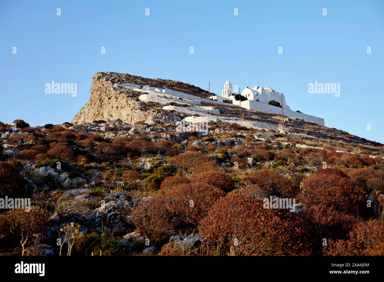 Weißes traditionelles Gebäude mit einem Glockenturm auf einem Hügel, umgeben von Sträuchern vor einem klaren blauen Himmel in der Abenddämmerung. Stockfoto