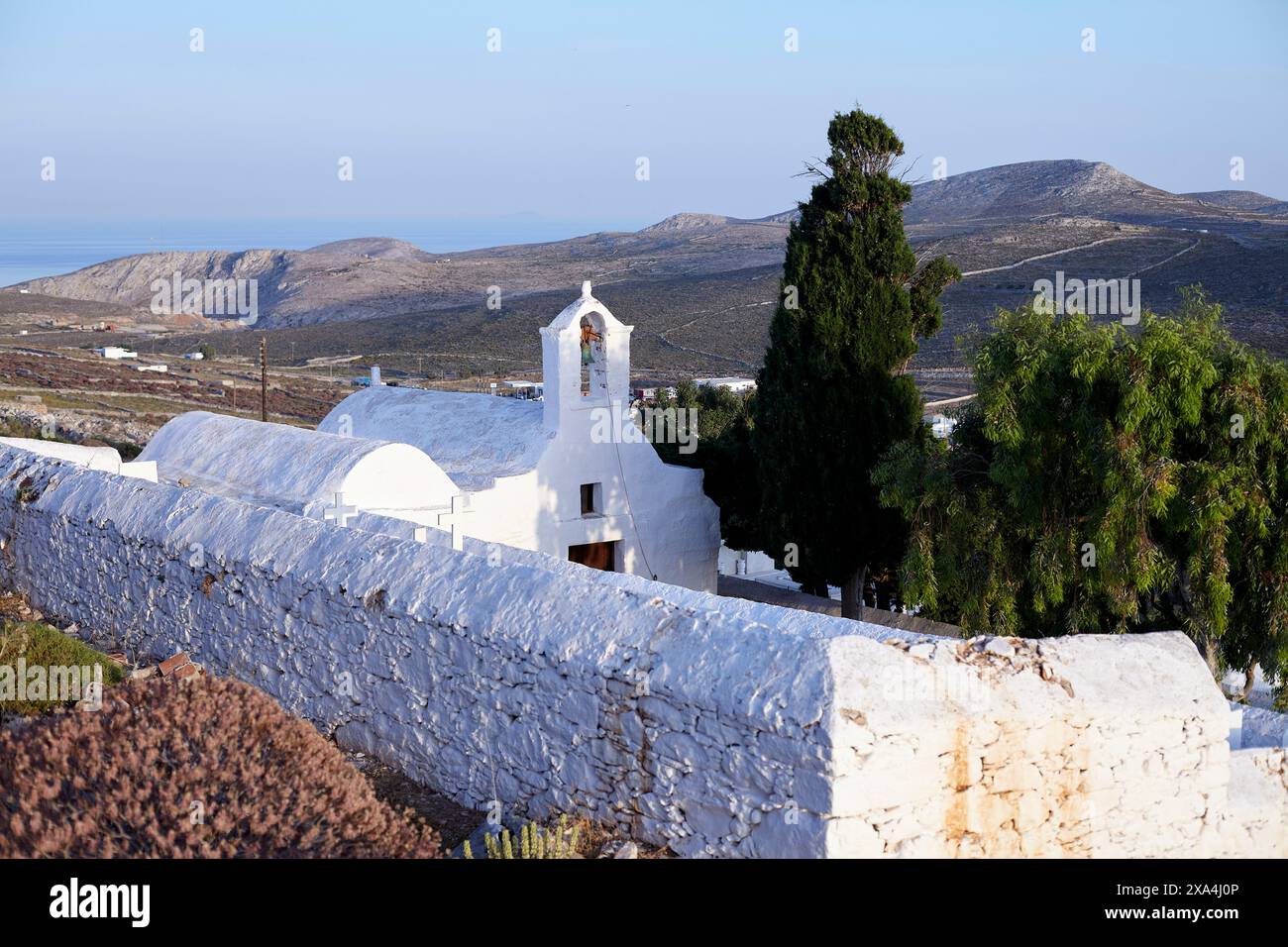 Eine ruhige Landschaft mit einer traditionellen weiß getünchten Kirche mit einem Glockenturm, umgeben von einer Steinmauer, mit Blick auf ein hügeliges Gelände mit dem Meer in der Ferne bei Sonnenaufgang. Stockfoto