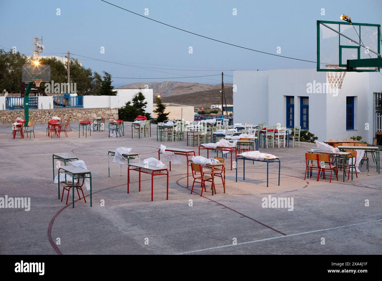 In der Abenddämmerung befindet sich ein Basketballfeld mit Tischen und Stühlen, die übersichtlich angeordnet sind, unter dem Licht der sanften Lichter, neben einem weißen Gebäude mit blauen Besätzen. Stockfoto