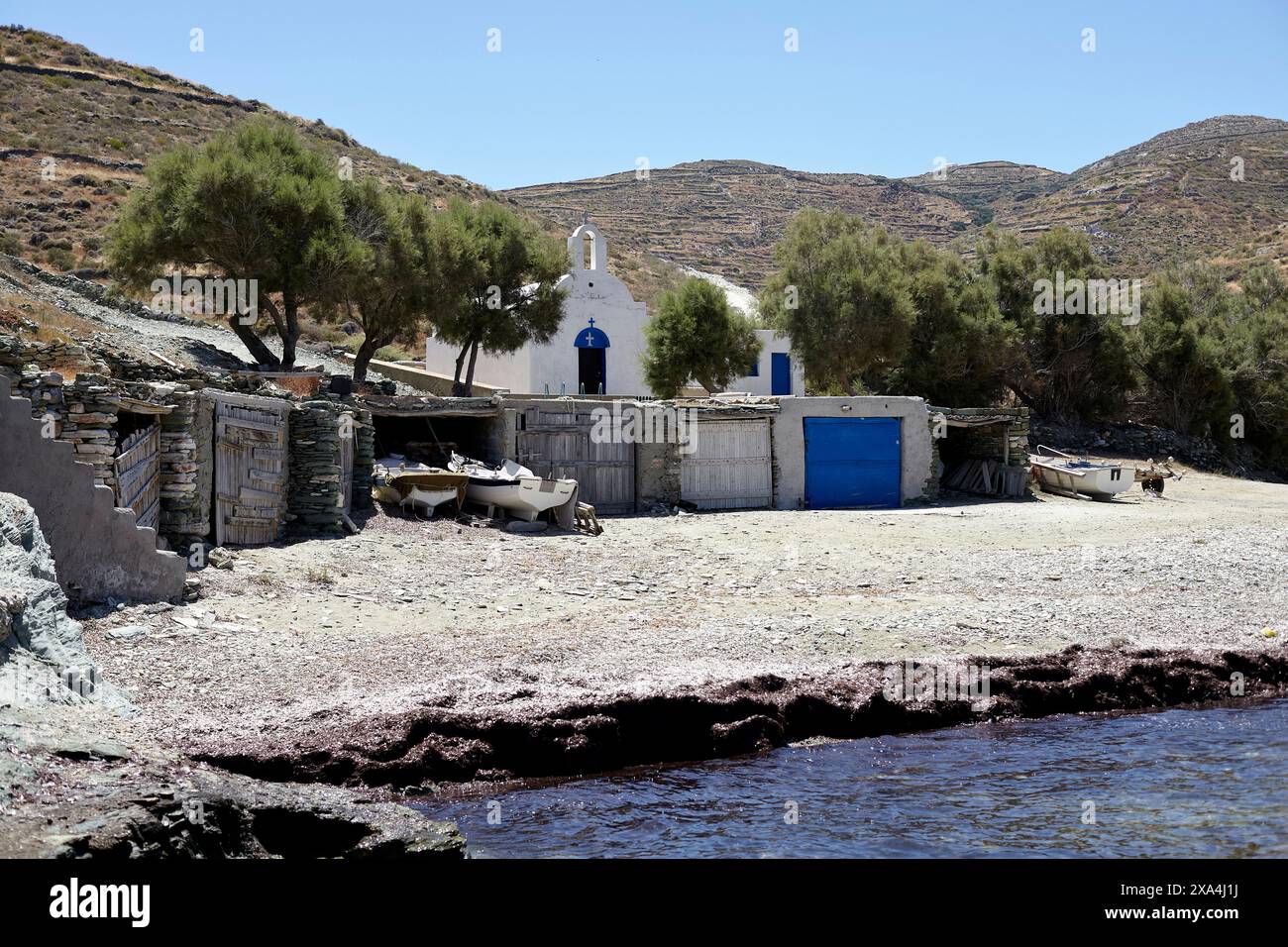Eine ruhige Küstenszene mit einer kleinen weißen Kirche mit einer blauen Kuppel vor einer hügeligen Landschaft mit umliegenden Sträuchern. Boote und Bauwerke befinden sich in der Nähe des Wasserrandes unter einem hellblauen Himmel. Stockfoto