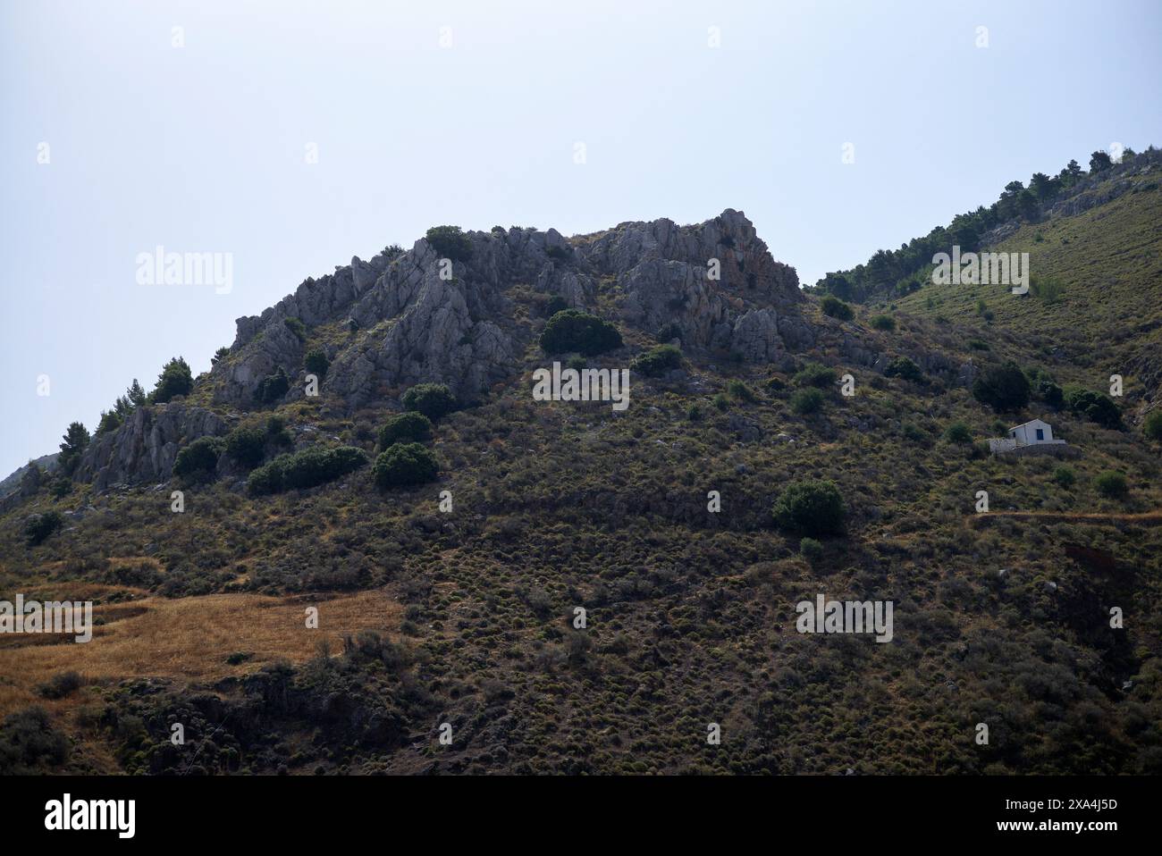 Ein zerklüfteter Hügel mit einem felsigen Ausläufer und einigen verstreuten Büschen unter klarem Himmel, mit einem kleinen weißen Gebäude unten rechts. Stockfoto