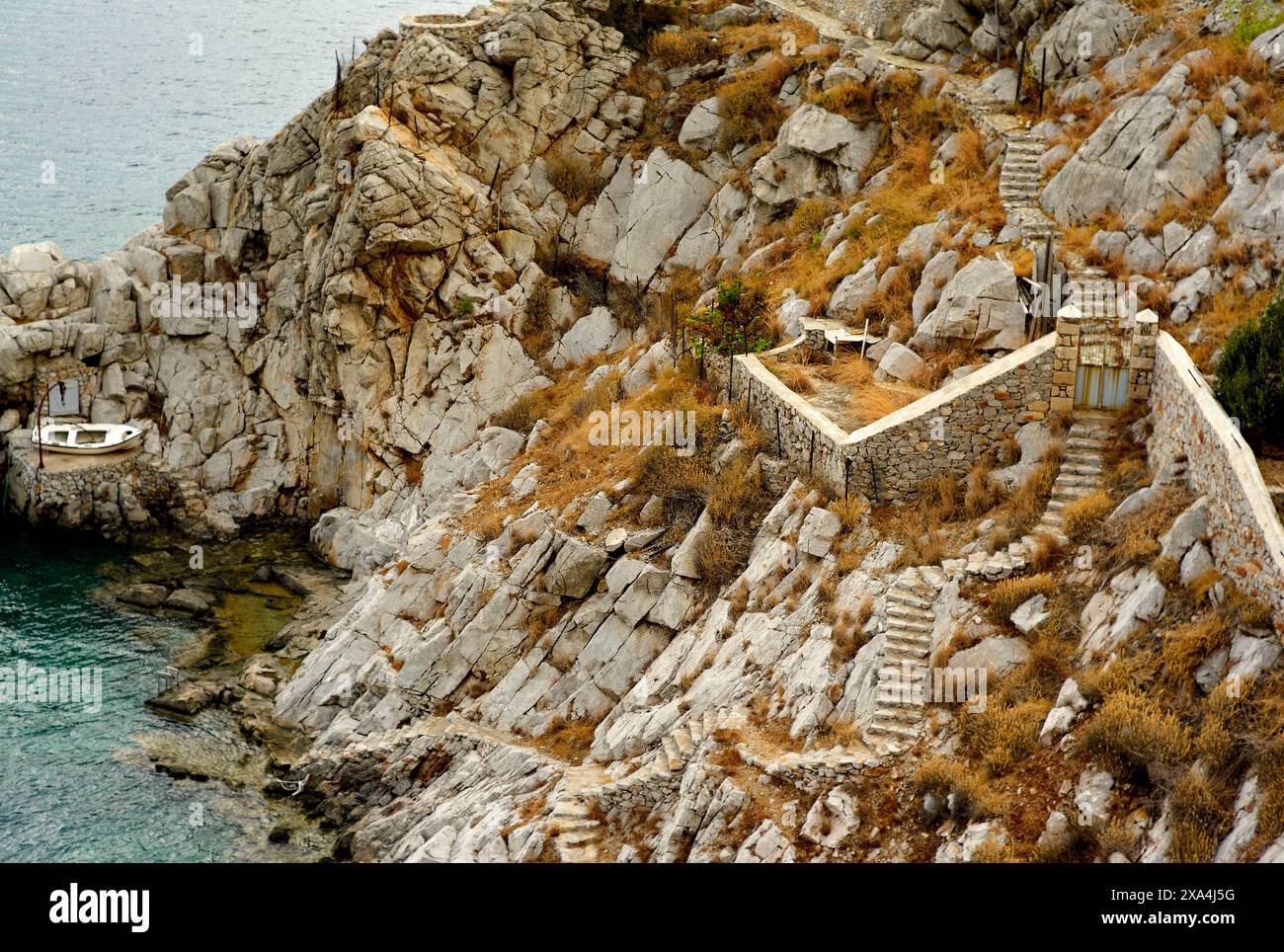 Eine Steintreppe führt über eine zerklüftete Klippe zu einem kleinen Dock, wo ein einsames Boot auf dem klaren blauen Wasser schwimmt. Stockfoto