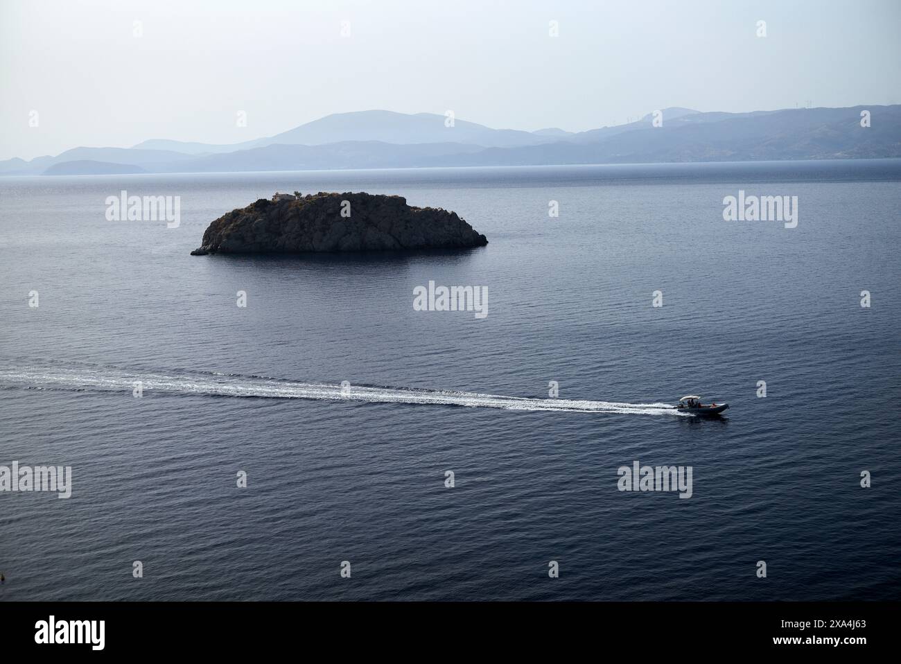 Ein Boot hinterlässt eine Spur auf dem ruhigen blauen Wasser, während es in Richtung einer kleinen felsigen Insel mit Bergen im fernen Hintergrund fährt. Stockfoto