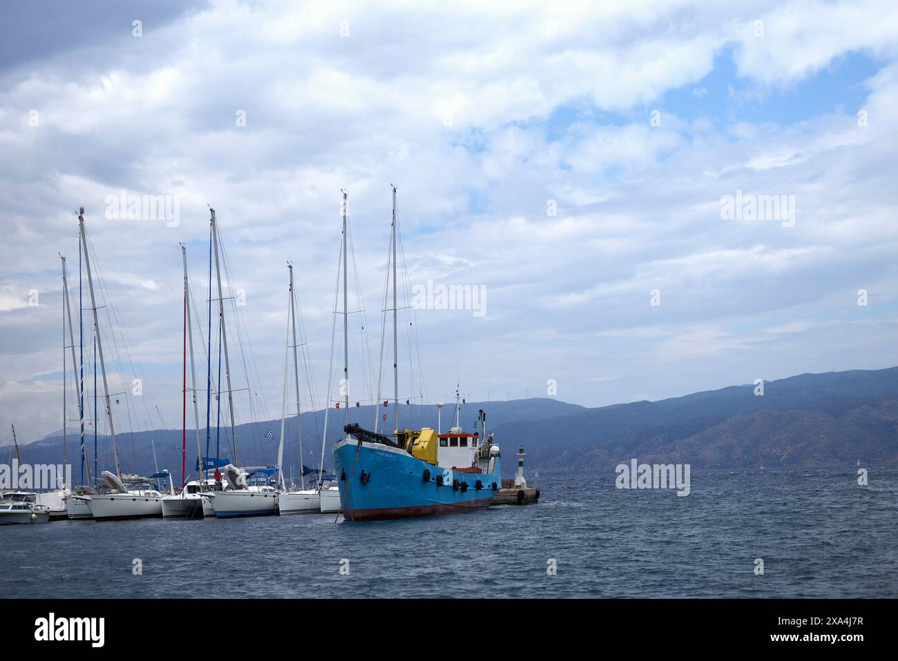 Ein blaues Fischerboot, das im Wasser verankert ist und von mehreren Segelbooten umgeben ist, vor dem Hintergrund des bewölkten Himmels und der fernen Berge. Stockfoto