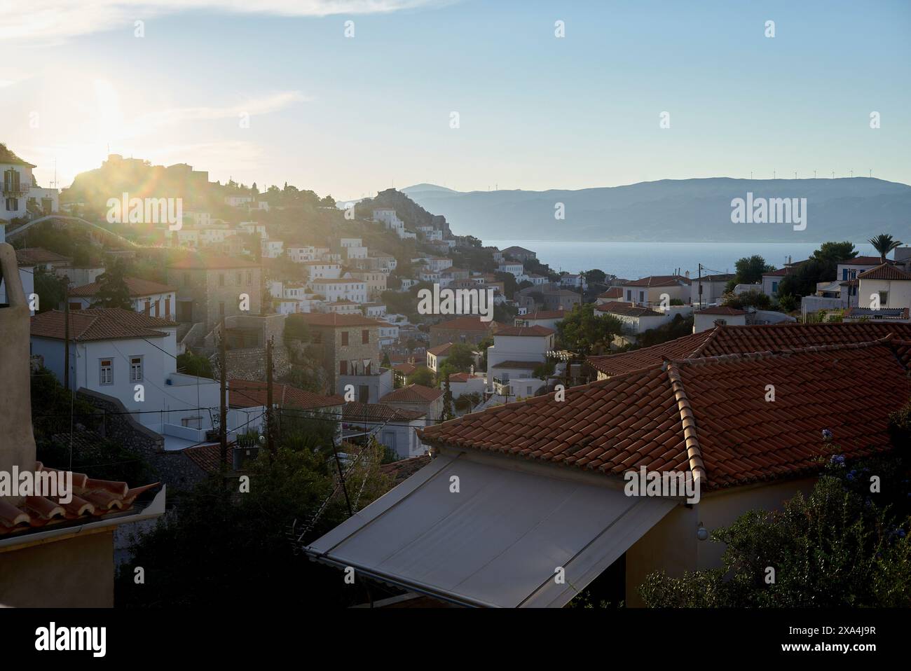 Ein malerischer Blick auf eine Küstenstadt bei Sonnenaufgang mit Terrakotta-Dächern und die Sonne, die über den Hügel blickt, wirft sanftes Licht auf die Gebäude und das ruhige Meer im Hintergrund. Stockfoto