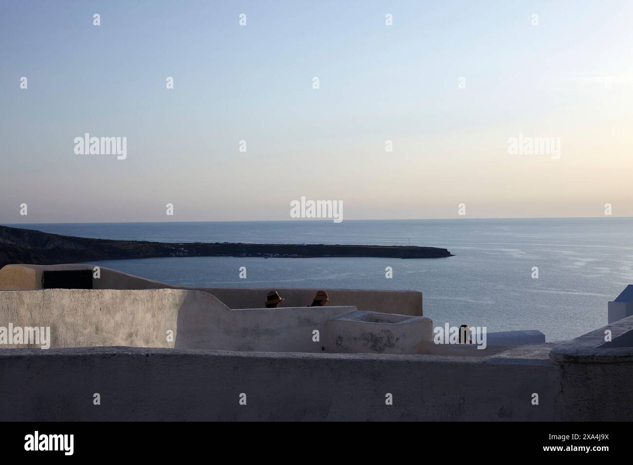 Silhouetten von Individuen sind über einer weiß getünchten Wand zu sehen, mit Blick auf ein ruhiges Meer in der Dämmerung, mit klarem Himmel und einem ruhigen Horizont. Stockfoto