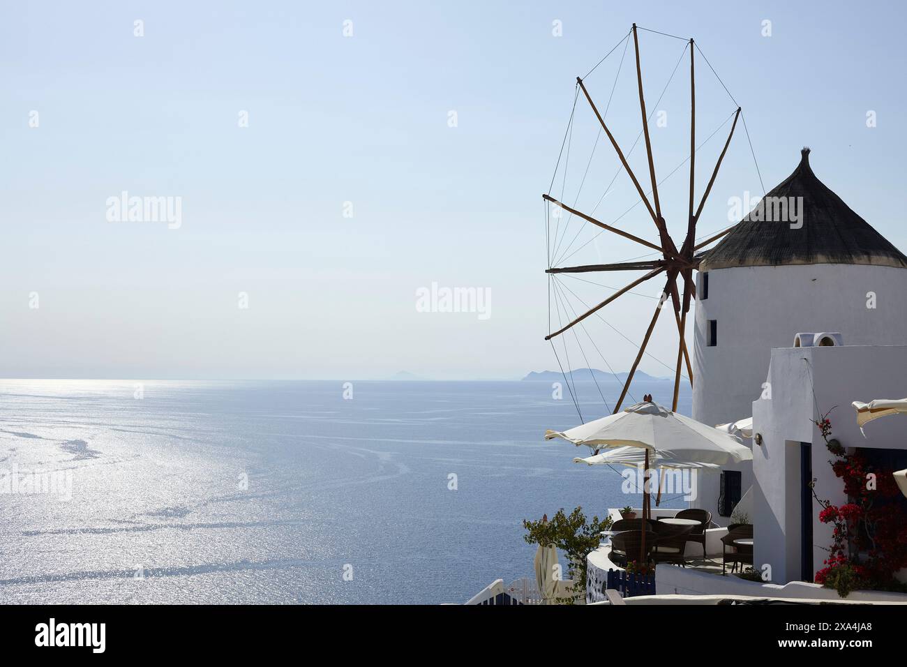 Eine traditionelle weiße Windmühle mit einem hölzernen Windrad überblickt das glitzernde blaue Meer von einem hohen Aussichtspunkt, mit einem klaren Himmel darüber und einem Hauch von einer fernen Insel am Horizont. Stockfoto