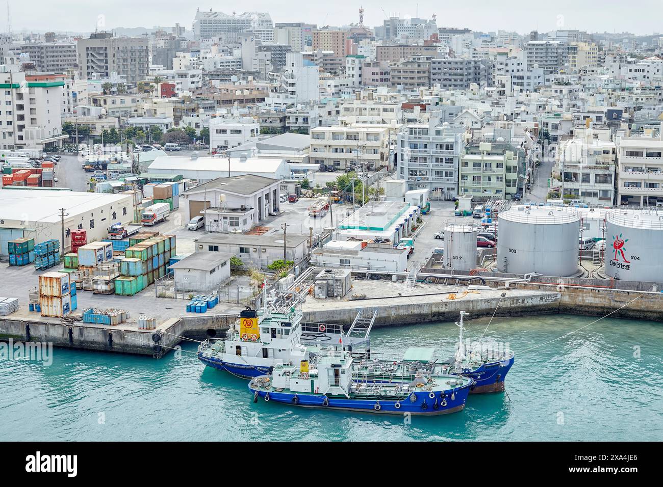 Blick aus der Vogelperspektive auf eine geschäftige Küstenstadt mit Schwerpunkt auf einem angedockten blauen Fischereischiff, umgeben von Industrieanlagen und Gebäuden. Stockfoto