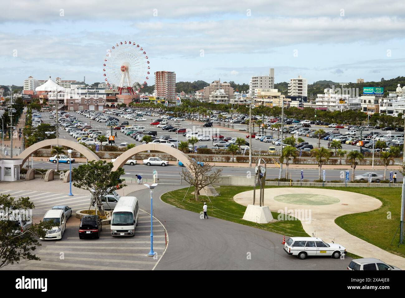 Ein Blick aus der Vogelperspektive auf einen belebten Parkplatz in einem Stadtgebiet mit einem großen Riesenrad in der Ferne, Gebäuden und einem klaren Himmel. Stockfoto