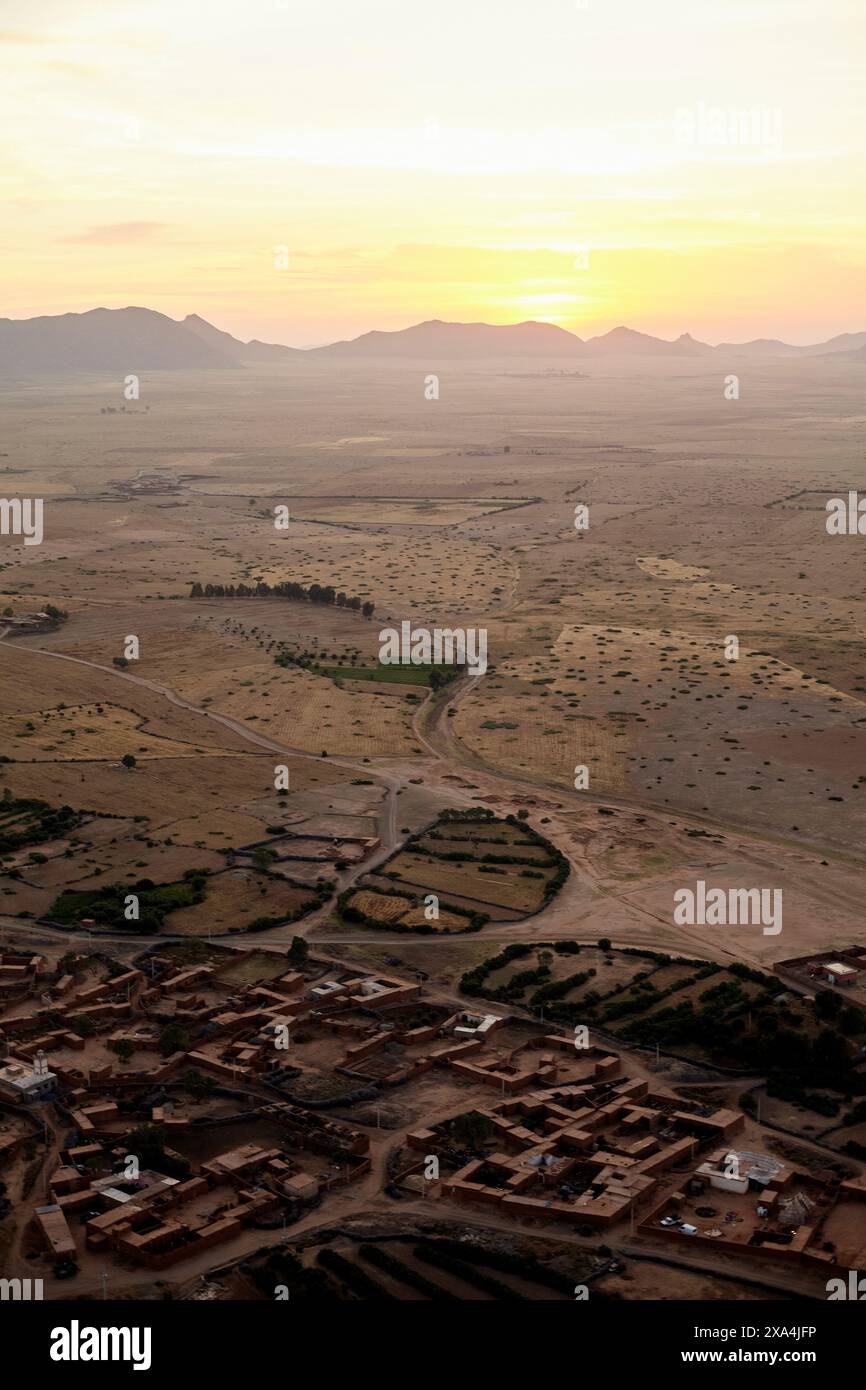 Ein Blick aus der Vogelperspektive auf eine Siedlung mit erdigen Gebäuden, umgeben von einer weiten, trockenen Landschaft bei Sonnenuntergang, mit Bergen in der Ferne und der Sonne, die unter dem Horizont eintaucht. Stockfoto