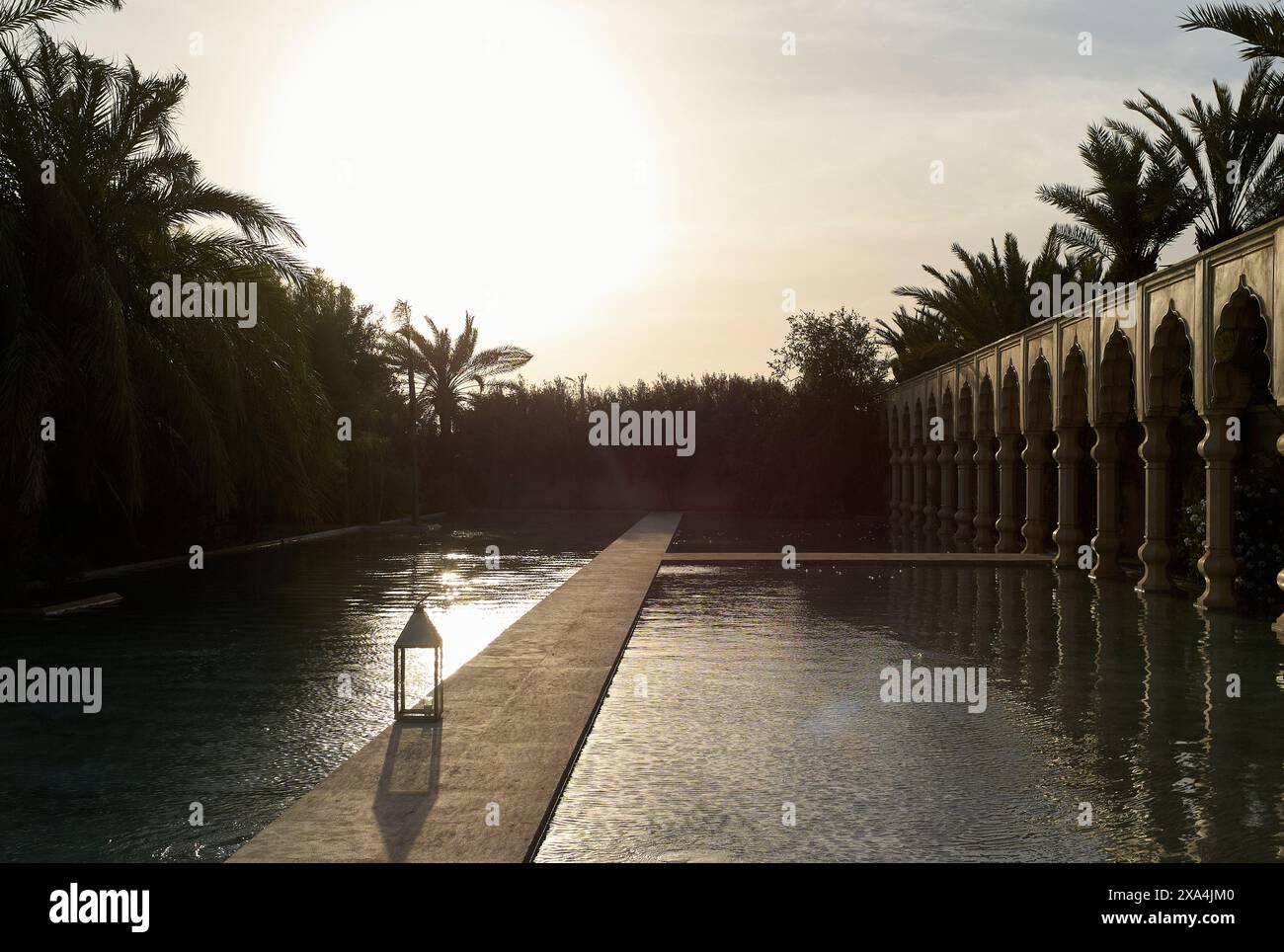 Das Bild zeigt einen ruhigen Swimmingpool in der Abenddämmerung, in dem das Sonnenlicht durch Palmen strömt und von der Wasseroberfläche reflektiert wird, neben einem kunstvollen Verzicht Stockfoto