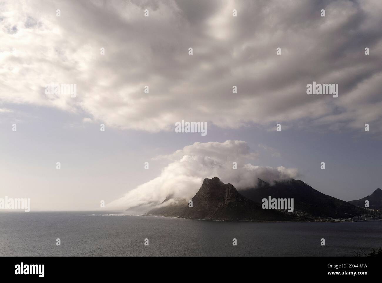 Wolken bedecken die zerklüfteten Gipfel eines Berges neben einem ruhigen Meer unter einem dramatischen Himmel. Stockfoto