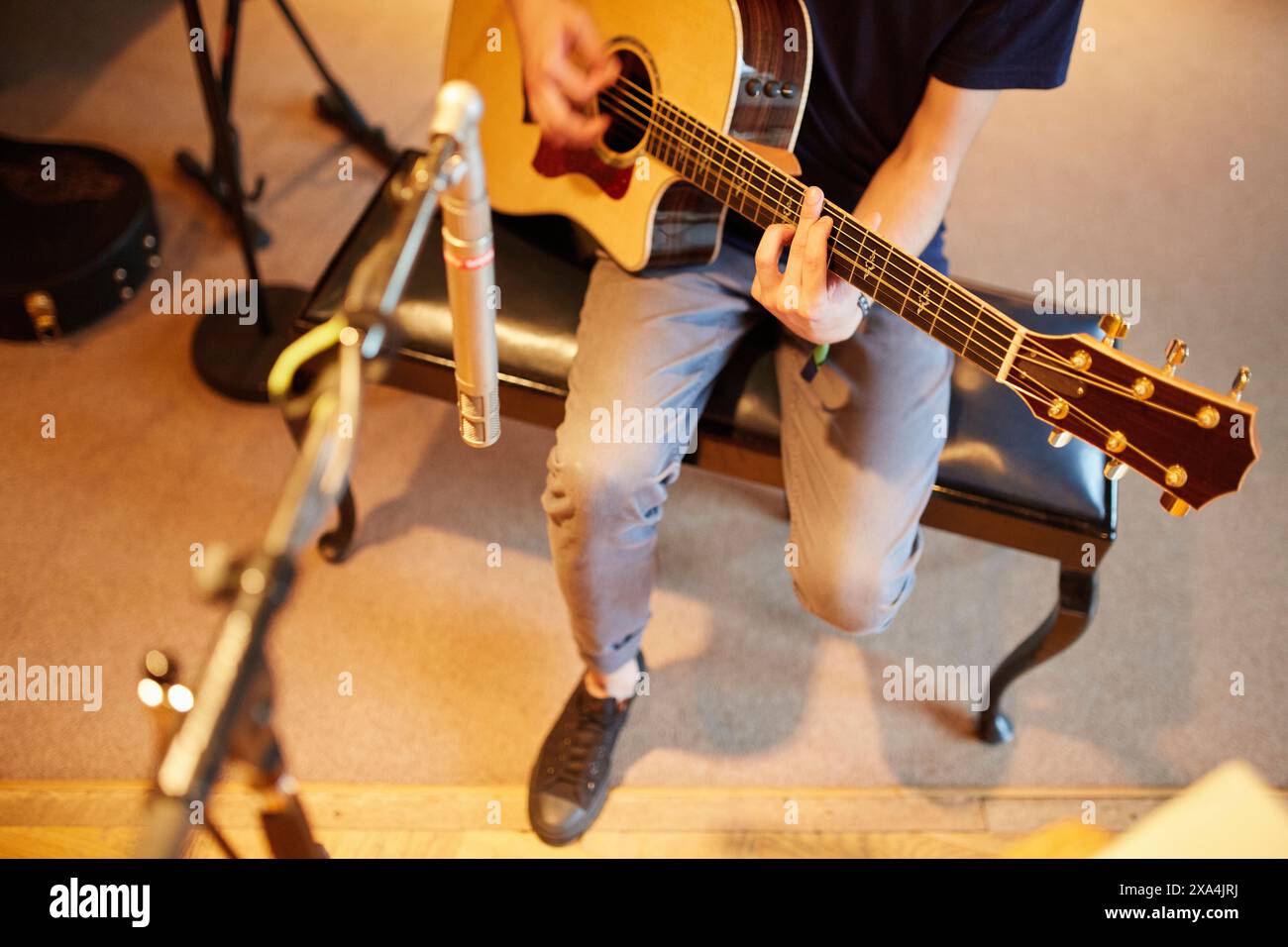 Ein Musiker sitzt auf einem Hocker und spielt eine Akustikgitarre in einem warm beleuchteten Raum mit einem Mikrofon und einer anderen Gitarre im Hintergrund. Stockfoto