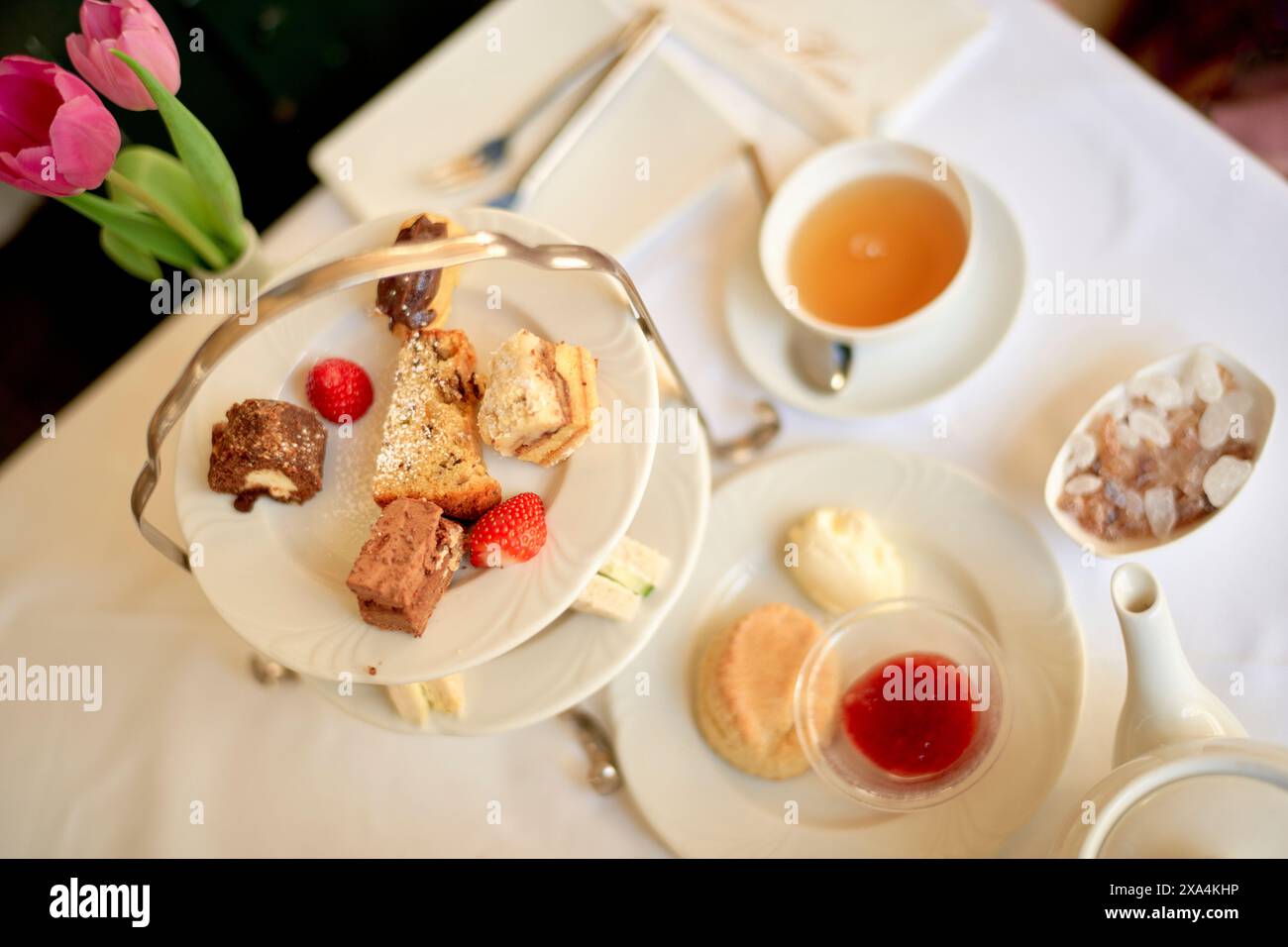 Ein eleganter Nachmittagstee mit einer Auswahl an Kuchen, einer Teetasse und einer Teekanne auf einem Tisch, die von einer Vase mit rosa Tulpen akzentuiert wird. Stockfoto