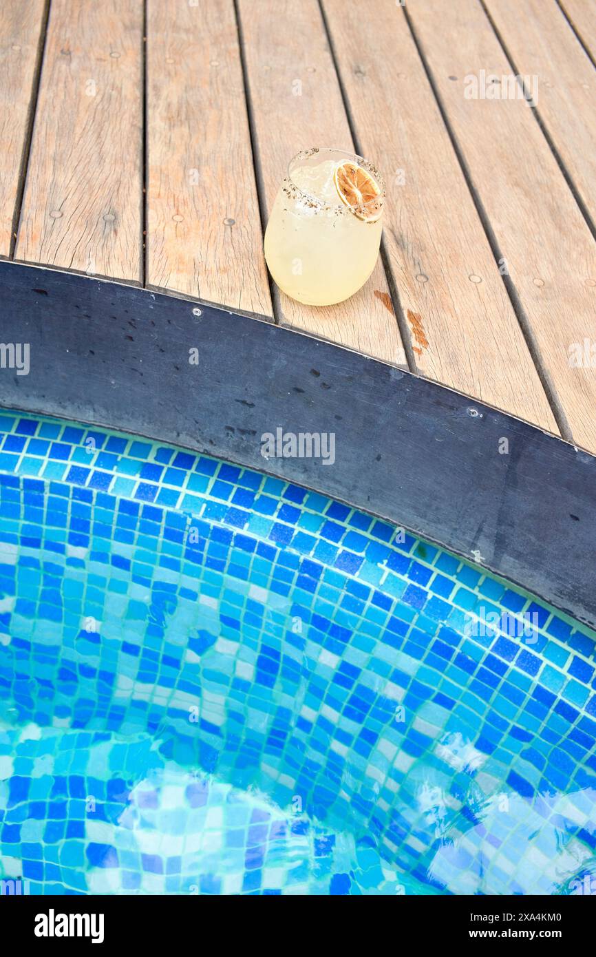Ein erfrischender Cocktail mit Zitrusgarnitur befindet sich auf einer Holzterrasse neben den leuchtend blauen Fliesen eines Swimmingpools. Stockfoto