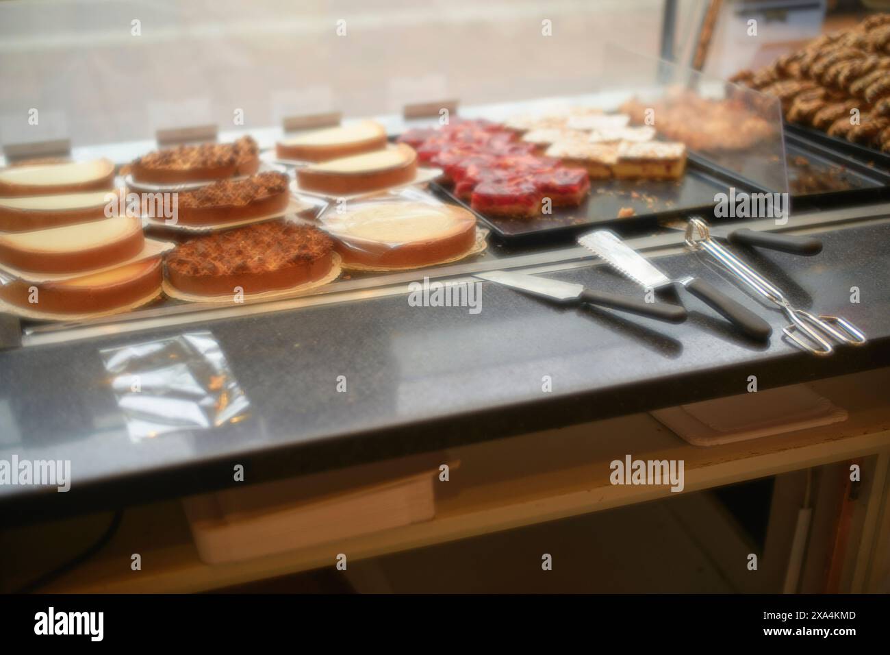 Eine Auswahl an Kuchen und Gebäck, die in einer Bäckerei hinter einer Glastheke präsentiert werden, mit Servierutensilien im Vordergrund. Stockfoto