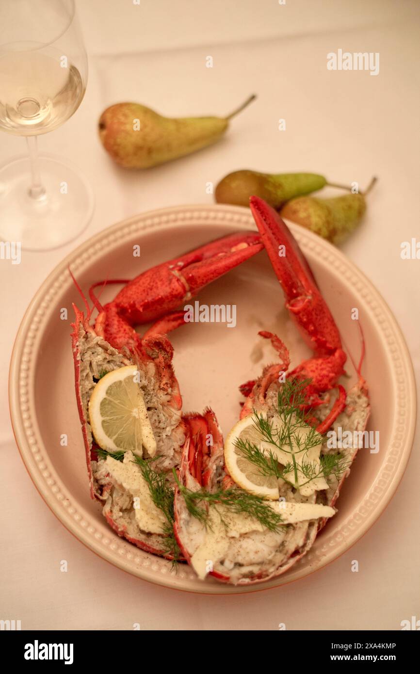 Ein überzogenes Meeresfrüchte-Gericht besteht aus einem gekochten Hummer, garniert mit Zitrone und Dill, begleitet von einem Glas Weißwein und zwei Birnen im Hintergrund, alles auf einem eleganten Tisch. Stockfoto