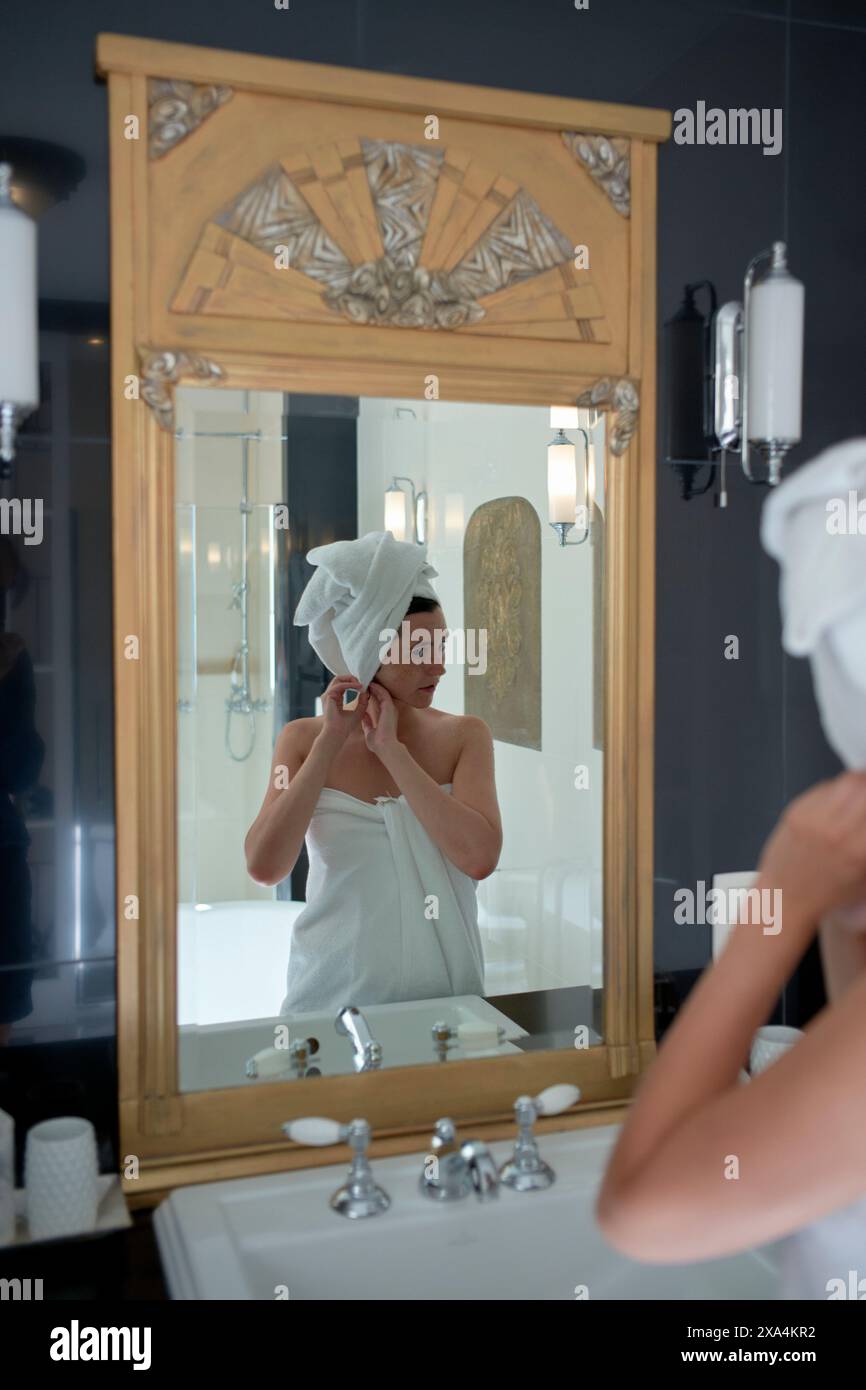 Eine Person, die in ein Handtuch gewickelt ist, steht in einem Badezimmer und passt einen Ohrring an, während sie ihre Reflexion in einem Spiegel betrachtet, der von einem kunstvollen Holzrahmen eingerahmt wird. Stockfoto
