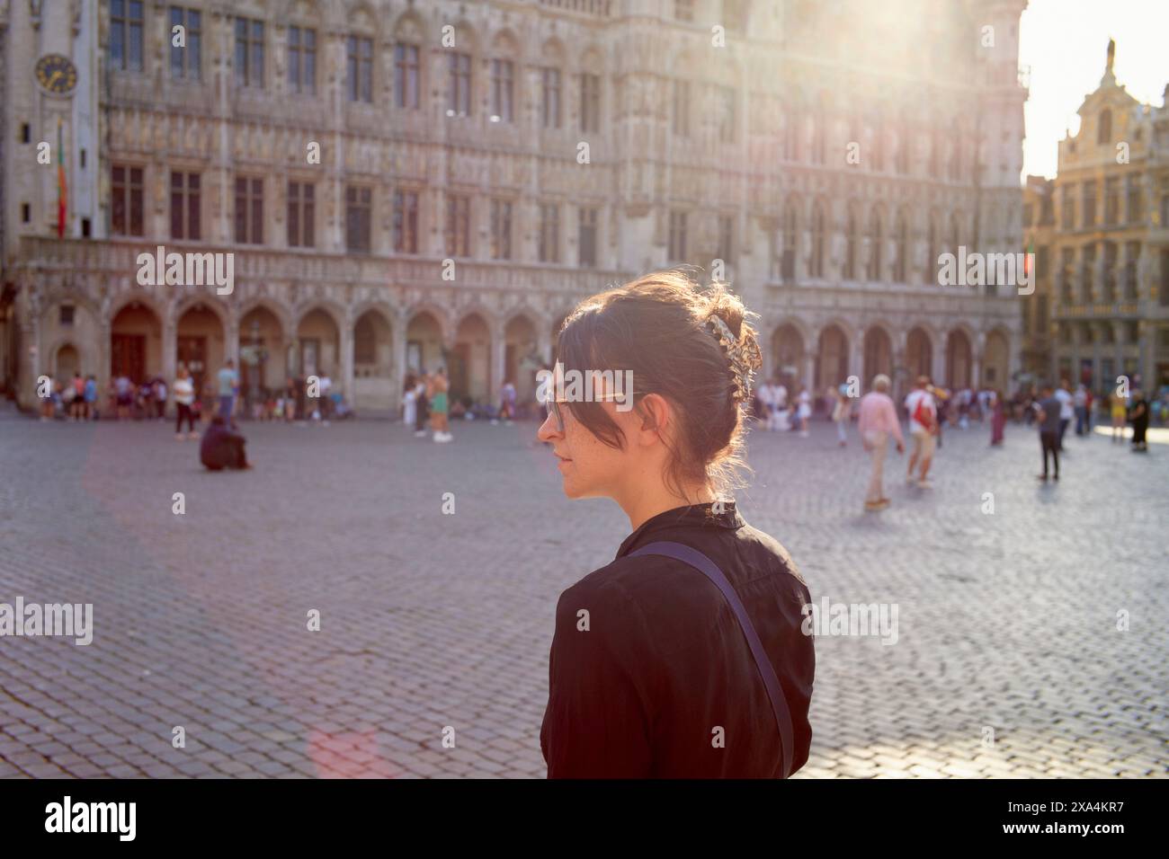 Eine Frau steht auf einem historischen Platz, die Sonne strahlt ein warmes Leuchten aus, während sie untergeht. Die Gebäude hinter ihr weisen komplizierte architektonische Details auf. Stockfoto