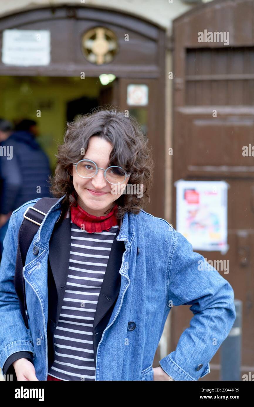Vor einer Holztür steht eine lächelnde Frau mit lockigem Haar, einer Jeansjacke, einem gestreiften Hemd und einem roten Schal. Stockfoto