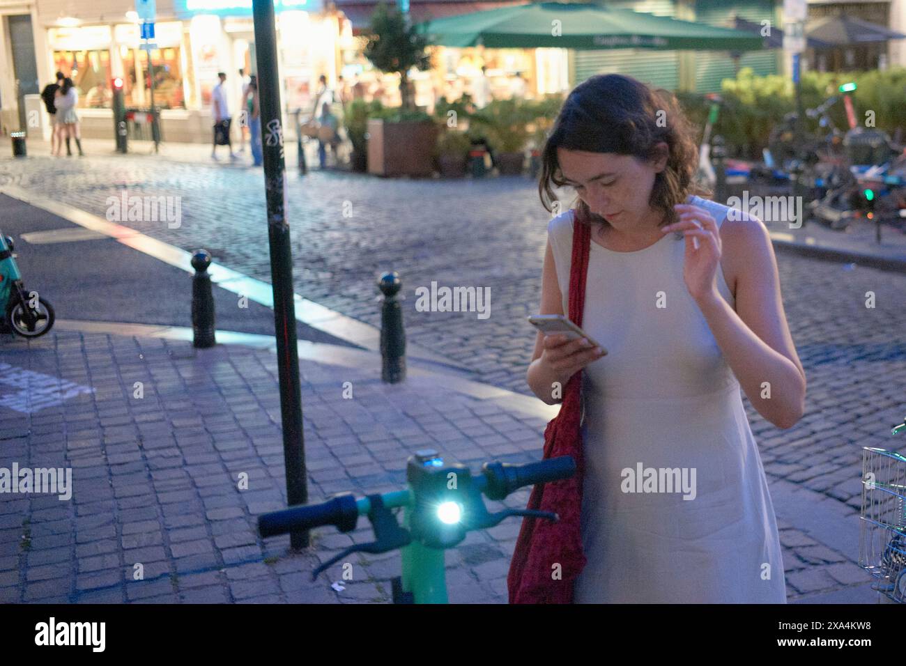 Eine Frau in einem weißen Kleid konzentriert sich auf ihr Smartphone in einer Stadtstraße in der Dämmerung, mit verschwommenen Hintergrundfiguren und Straßenlaternen. Stockfoto