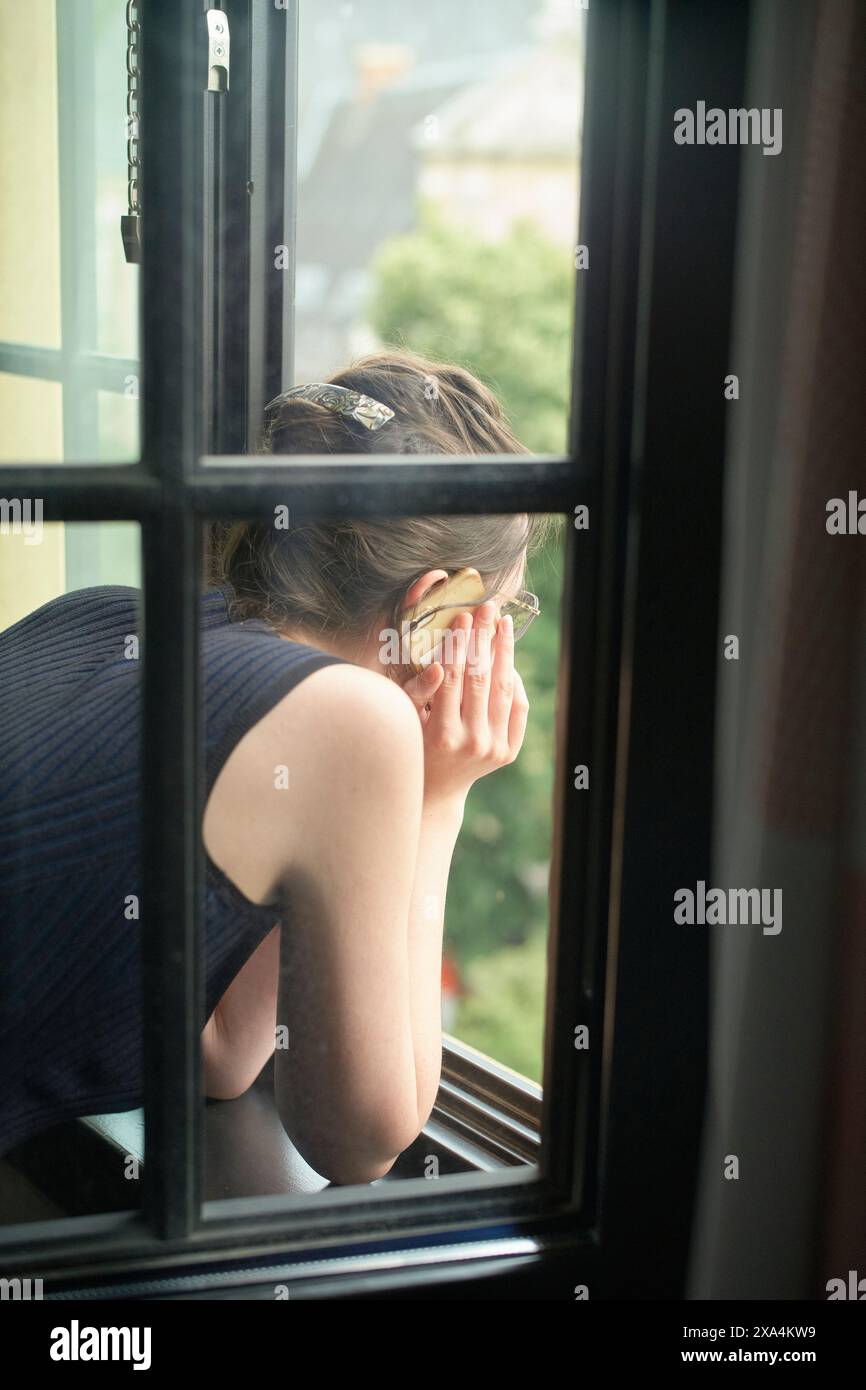 Eine Person wird von hinten gesehen, das Kinn auf der Hand aufliegt und aus einer Fensterscheibe blickt und ein Smartphone am Ohr hält. Stockfoto