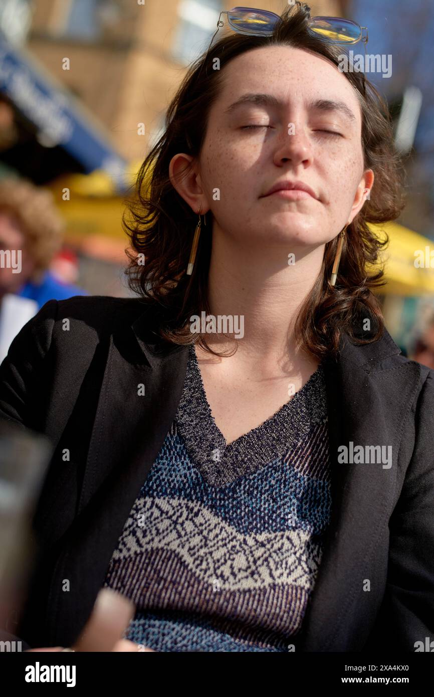 Ein junger Erwachsener mit geschlossenen Augen und einem entspannten Ausdruck saugt die Sonne auf, trägt ein gemustertes Oberteil, eine schwarze Jacke und eine Sonnenbrille auf den Kopf. Stockfoto