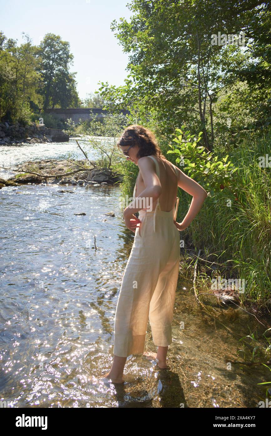 Eine Frau in hellem Kleid steht an einem Fluss und passt die Rückseite ihres Kleides in einer ruhigen natürlichen Umgebung mit Bäumen und einer Brücke im Hintergrund an. Stockfoto