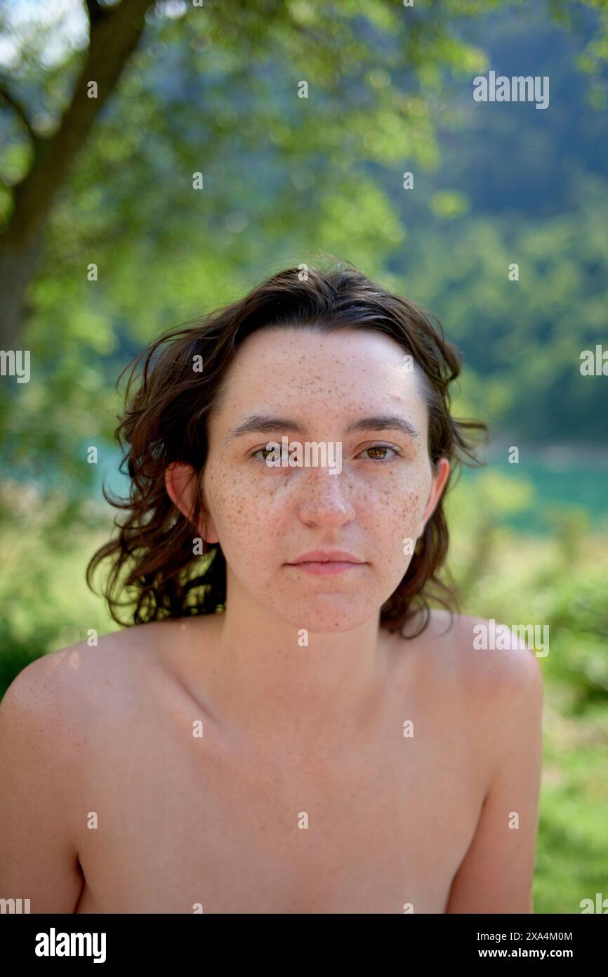 Ein Porträt einer jungen Frau mit Sommersprossen, braunem Haar und neutralem Ausdruck, die draußen mit einem unscharfen natürlichen Hintergrund steht. Stockfoto