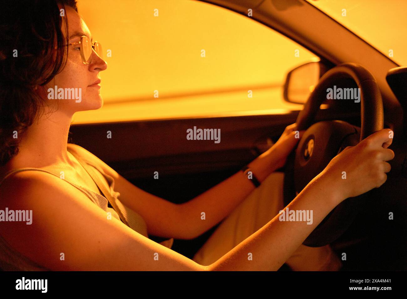 Eine Frau sitzt auf dem Fahrersitz eines Autos, Hände am Lenkrad, mit einem fokussierten Blick auf ihrem Gesicht, beleuchtet von einem warmen, orangen Licht. Stockfoto