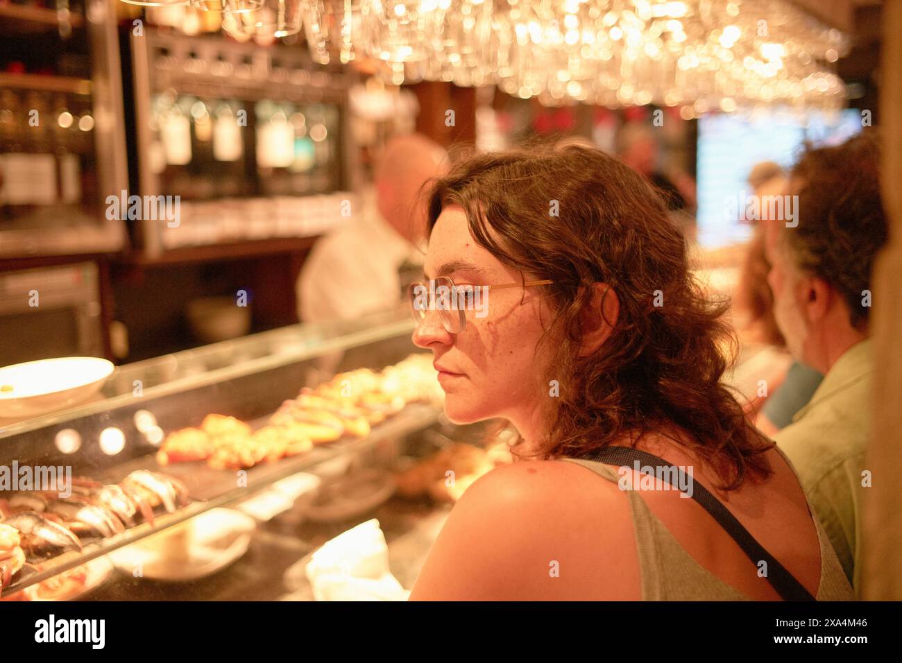 Eine Frau sitzt in einer Bar mit einem nachdenklichen Ausdruck und blickt auf ihre Seite, während die warme Umgebungsbeleuchtung eine Reihe von Tapas im Hintergrund beleuchtet. Stockfoto