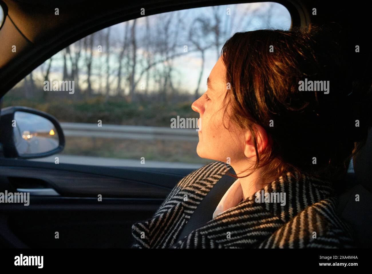 Eine Frau sitzt in einem Auto und schaut während eines Sonnenuntergangs durch das Beifahrerfenster. Sanftes Sonnenlicht beleuchtet die Hälfte ihres Gesichts. Stockfoto
