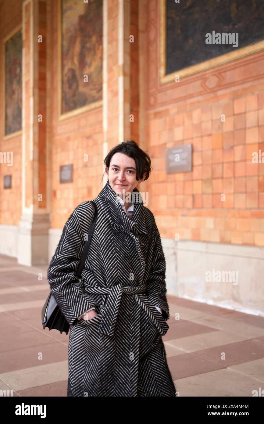 Eine Person steht vor einer reich verzierten Wand mit Wandmalereien, trägt einen Hahnentrittmantel und trägt eine schwarze Tasche. Stockfoto