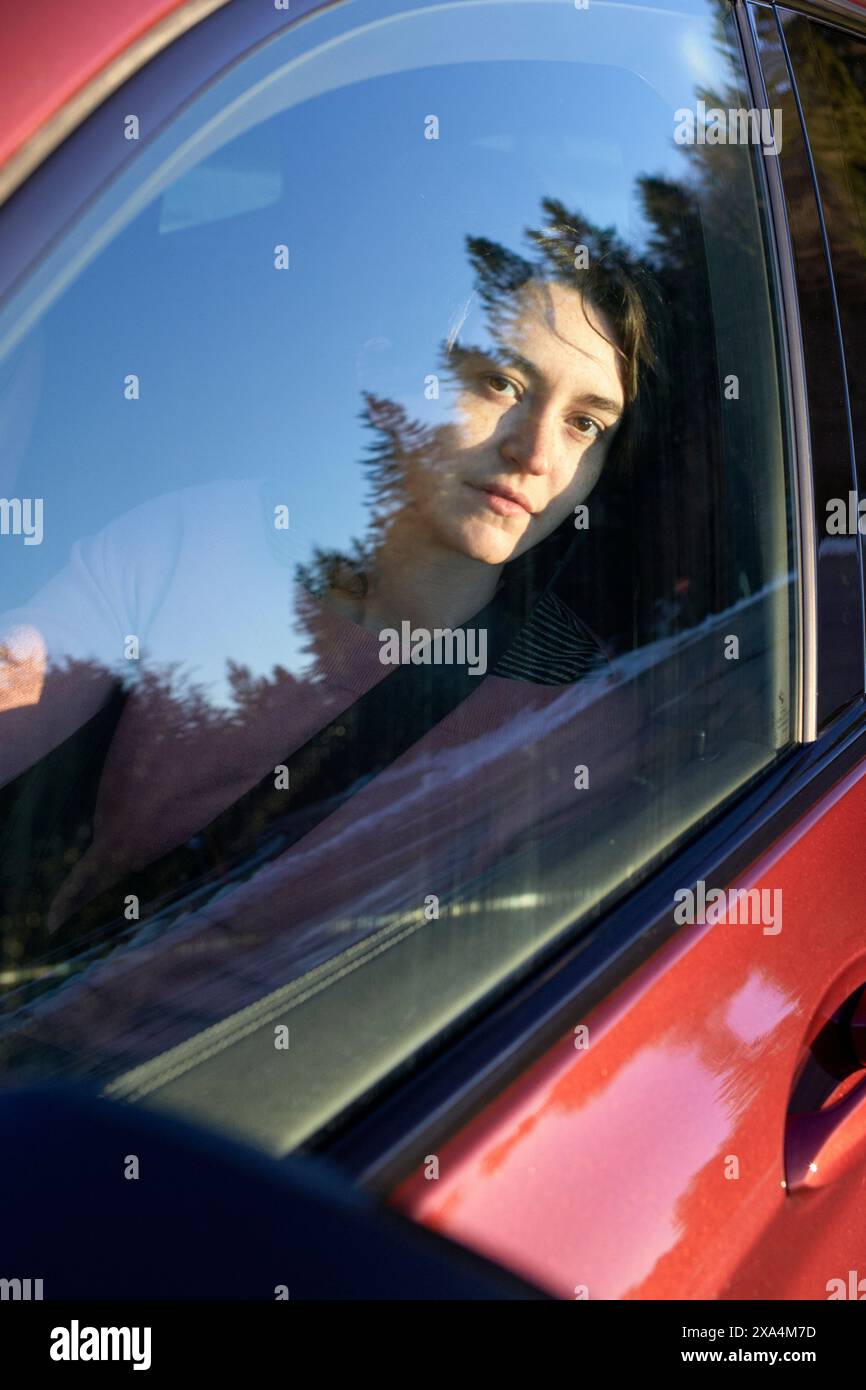 Eine Person schaut durch ein Autofenster, wobei ihr Gesicht teilweise durch die Reflektionen von Bäumen verdeckt ist. Stockfoto