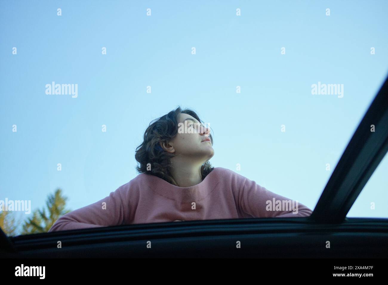 Eine Person blickt mit einem nachdenklichen Ausdruck nach oben, während ihr Kopf und ihre Schultern vor einem klaren blauen Himmel vom Schiebedach eines Autos auftauchen. Stockfoto