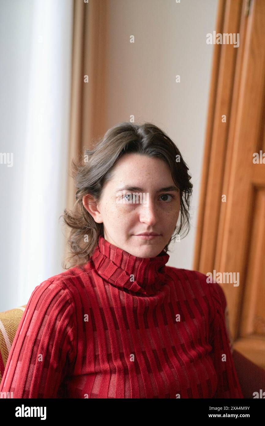 Eine Frau in einem roten Rollkragenpullover sitzt vor einem cremefarbenen Vorhang und neben einer Holztür und blickt neutral in die Kamera. Stockfoto