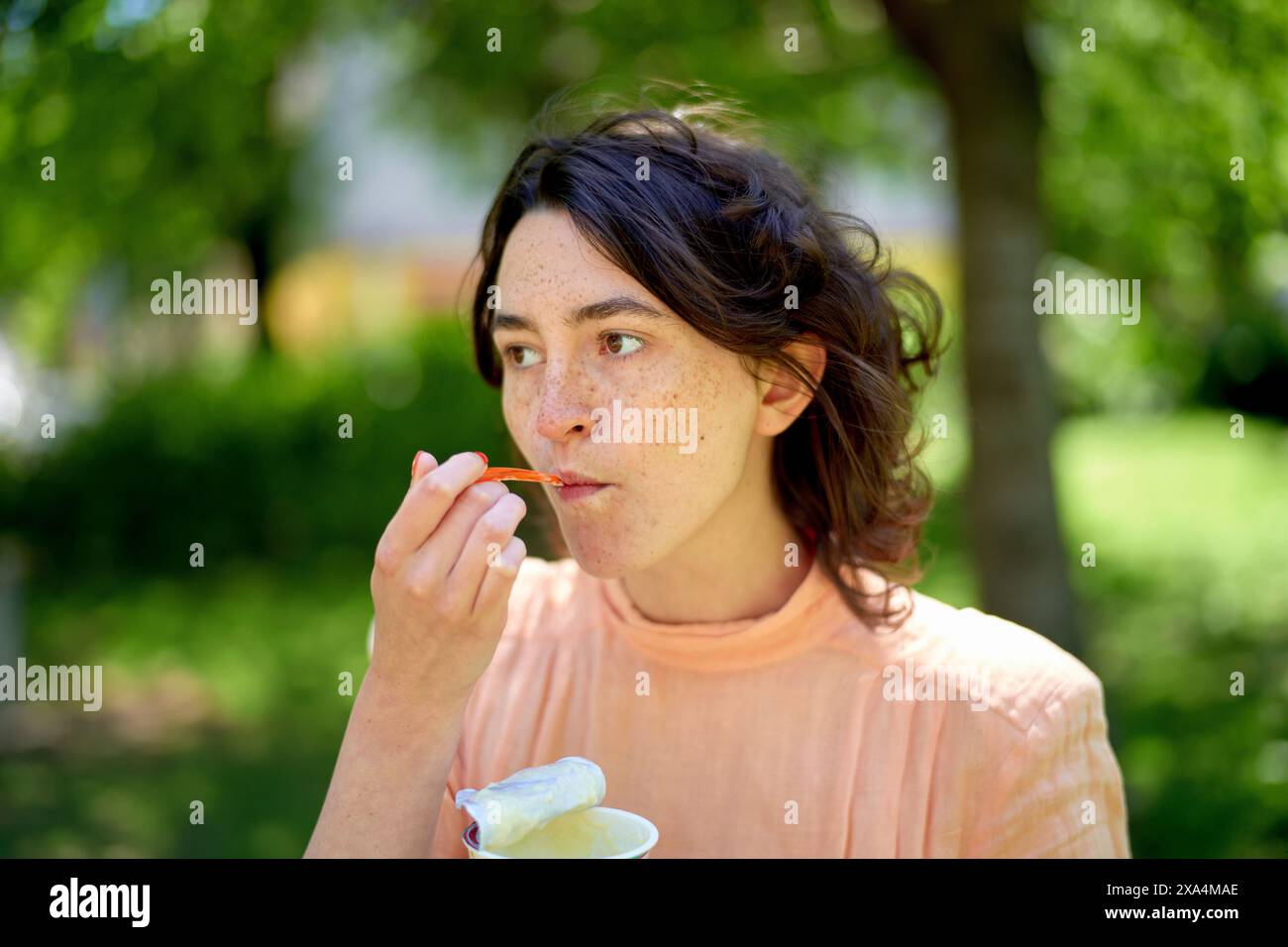 Eine junge Frau mit Sommersprossen isst draußen mit einem Plastiklöffel, mit Bäumen und einem Gebäude, das sanft im Hintergrund verschwimmt. Stockfoto
