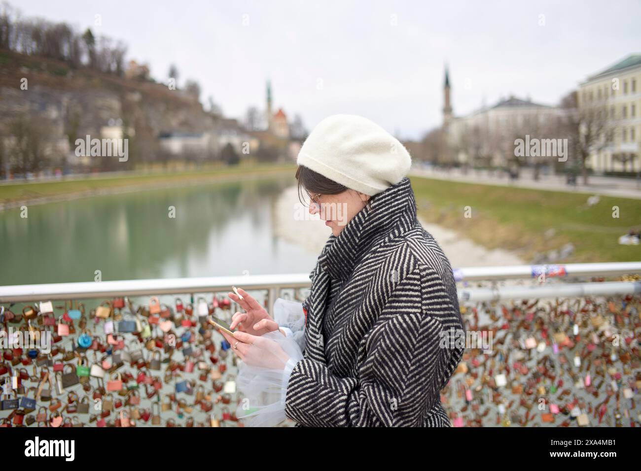 Eine Person steht auf einer Brücke mit Blick auf einen Fluss mit zahlreichen Liebesschlössern am Geländer, während sie ihr Smartphone überprüft. Stockfoto