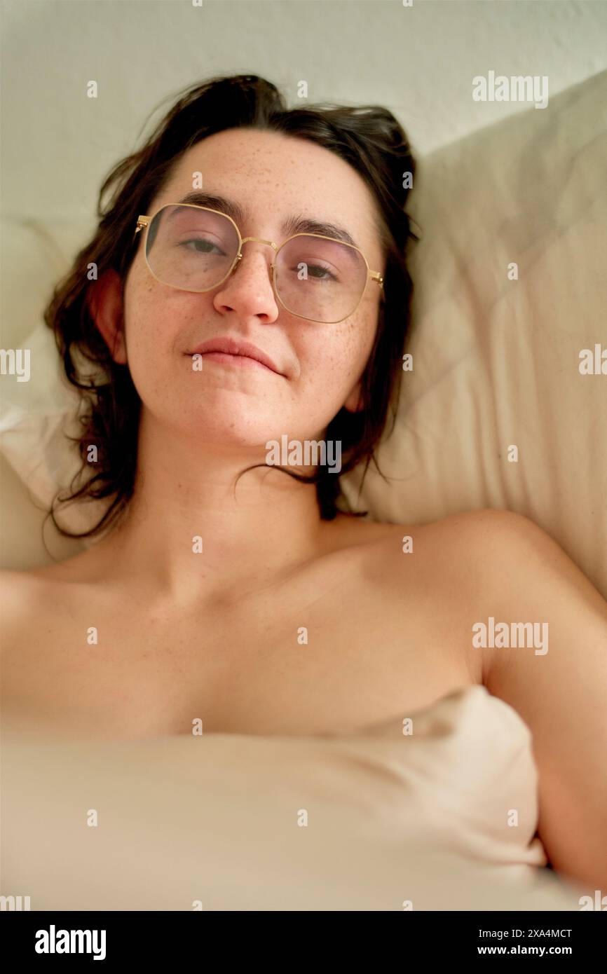 Ein junger Erwachsener mit Brille liegt hin und gibt einen entspannten Ausdruck mit einem subtilen Lächeln. Der Kopf liegt auf einem Kissen. Stockfoto