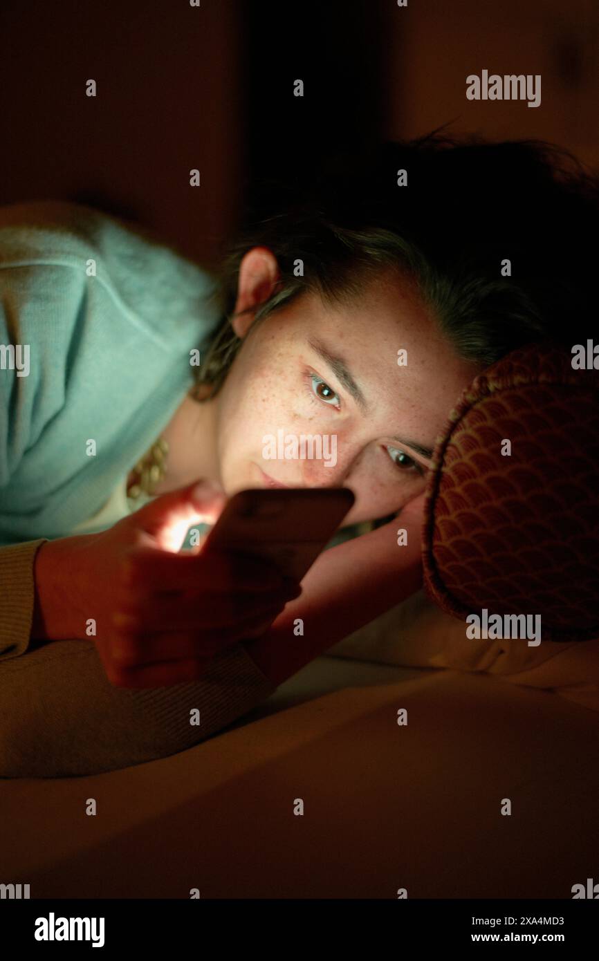 Eine Person liegt im Bett, beleuchtet durch das Leuchten eines Smartphones in einem dunklen Raum, deren Aufmerksamkeit vom Gerät gefangen wird. Stockfoto