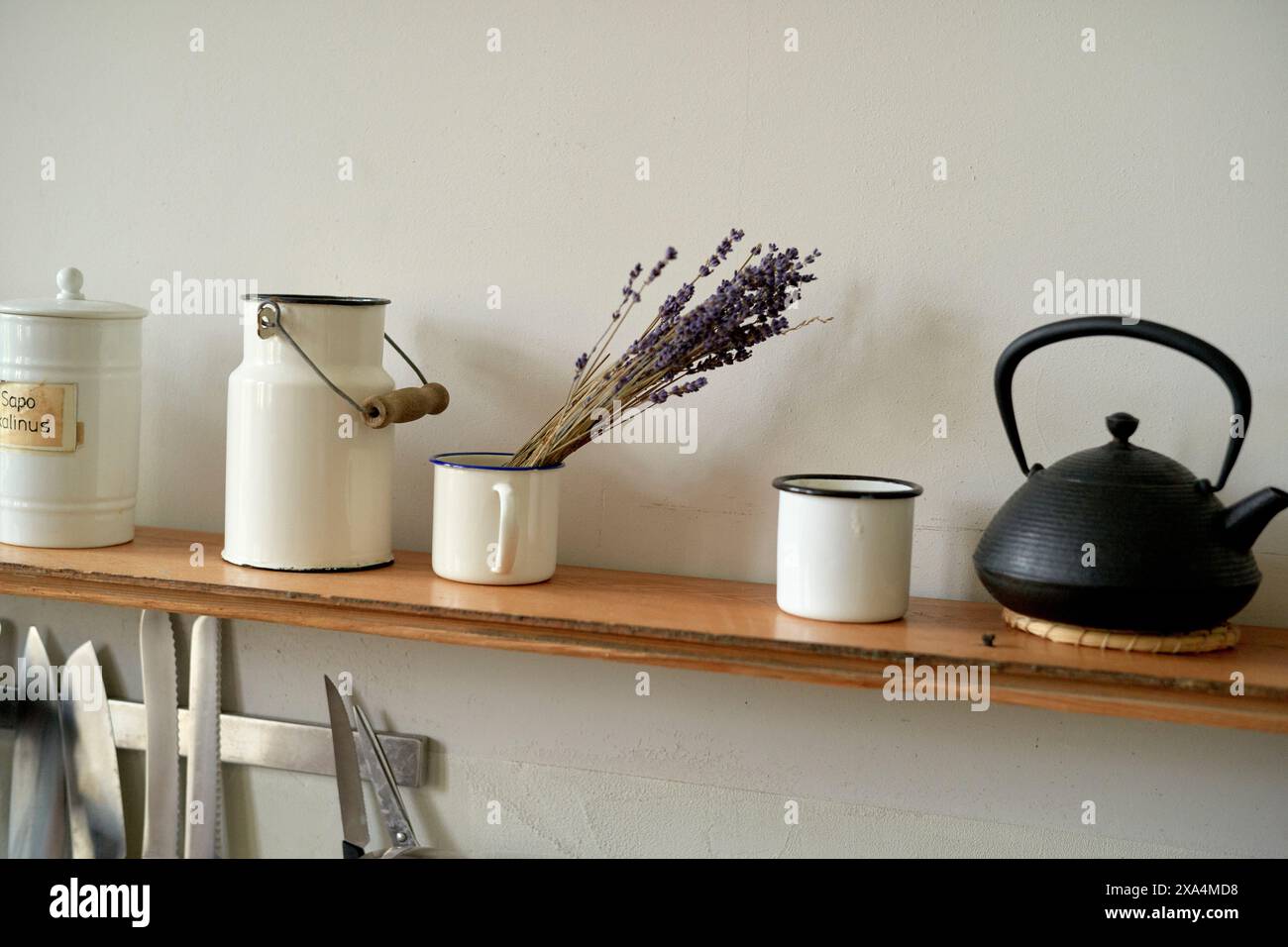 Ein Küchenregal mit einer Vintage-Emailleware-Sammlung, darunter eine weiße Kaffeekanne, eine Tasse mit getrocknetem Lavendel, eine kleine Schüssel und eine schwarze Teekanne, vor einer neutralen Wand. Stockfoto