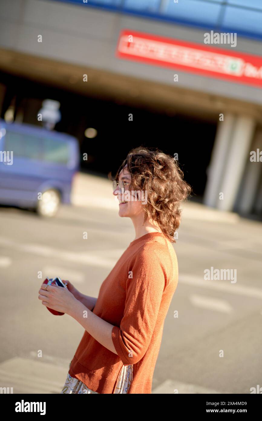 Eine Person mit lockigen Haaren lächelt und steht in einem sonnendurchfluteten Bereich, möglicherweise auf einem Parkplatz, mit einem klaren blauen Himmel darüber und einem Gebäude mit rotem Text im Hintergrund. Stockfoto
