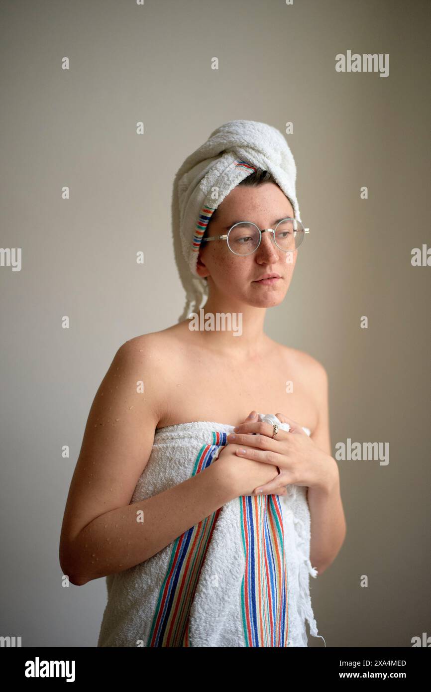 Eine junge Frau, die in ein Handtuch gewickelt ist und auf einem neutralen Hintergrund steht, eine Brille trägt, und ein anderes Handtuch um ihr Haar gewickelt. Stockfoto