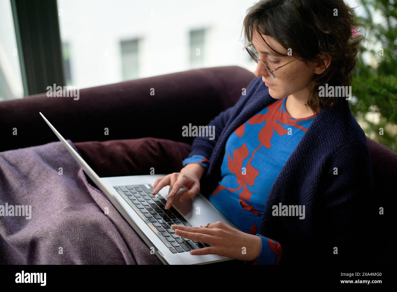 Eine Frau sitzt auf einer violetten Couch mit einem Laptop. Sie scheint sich auf ihre Arbeit konzentriert zu haben, wobei natürliches Licht den Raum von einem Fenster neben ihr beleuchtet. Stockfoto