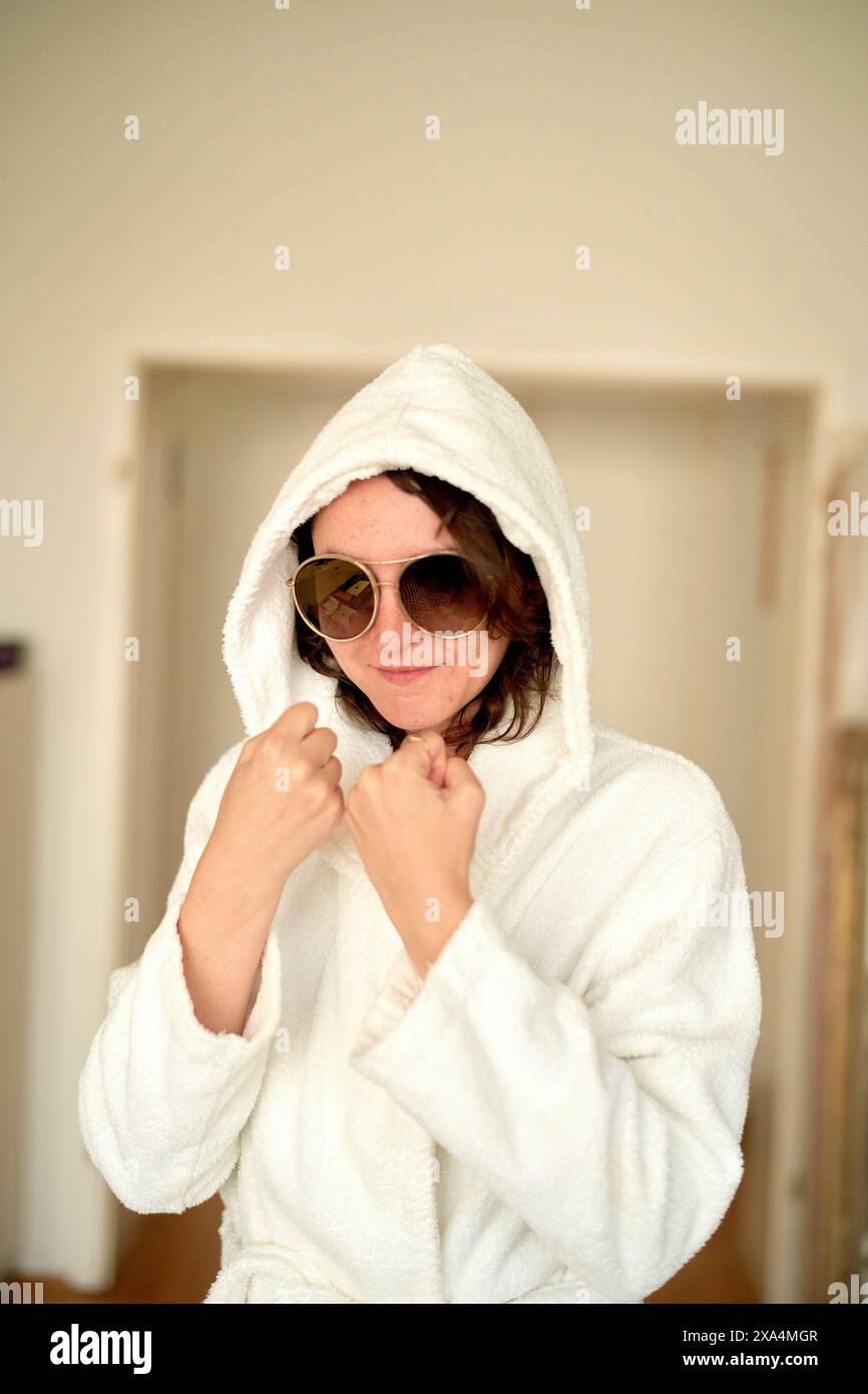 Eine Person trägt einen weißen Bademantel mit Kapuze und trägt eine dunkle Sonnenbrille und steht mit einer verspielten Pose drinnen. Stockfoto