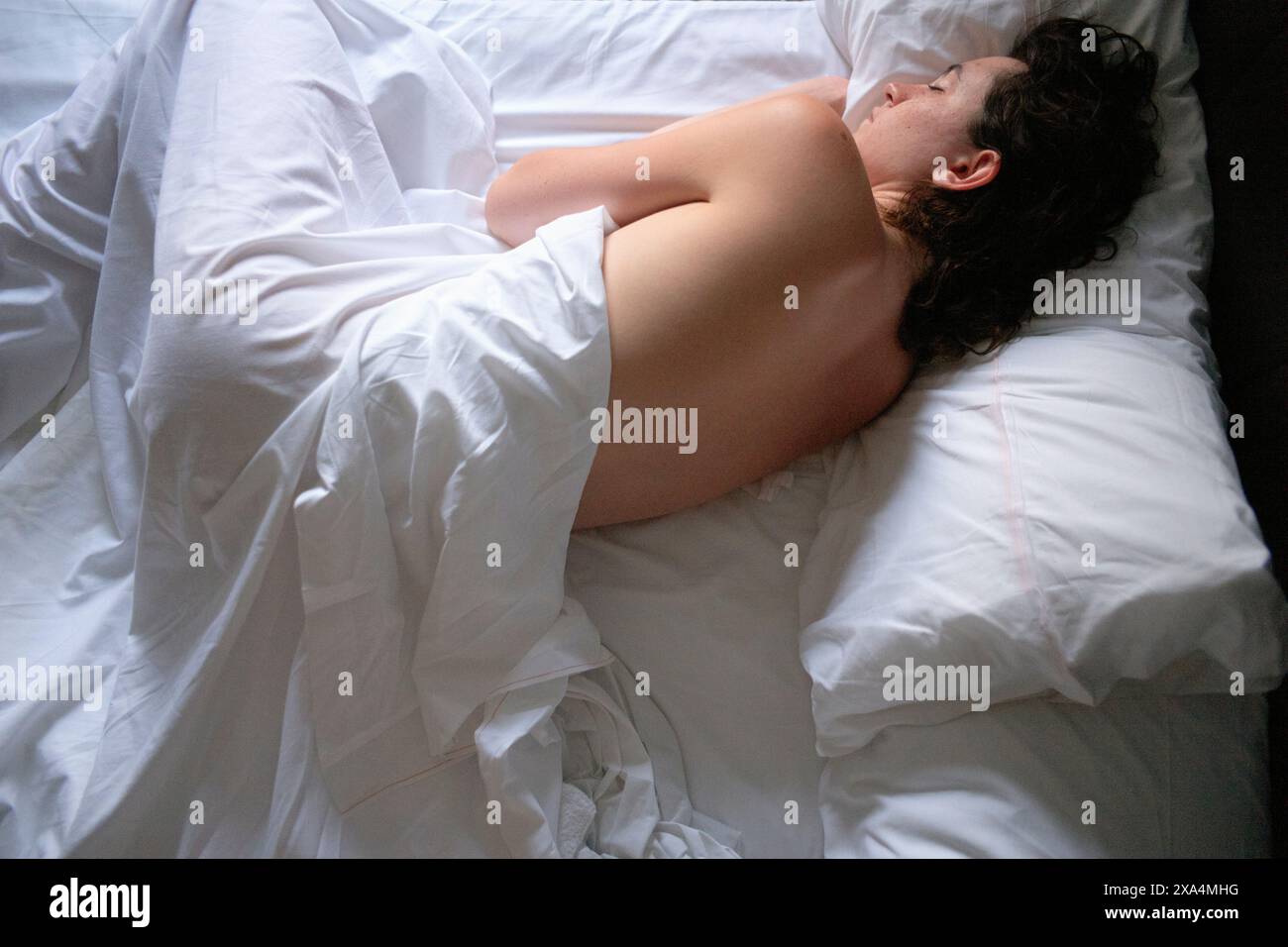 Eine junge Frau schläft friedlich auf einem Bett, das mit weißen Laken bedeckt ist, mit teilweise freiliegendem Rücken und Kopf auf einem weißen Kissen. Stockfoto