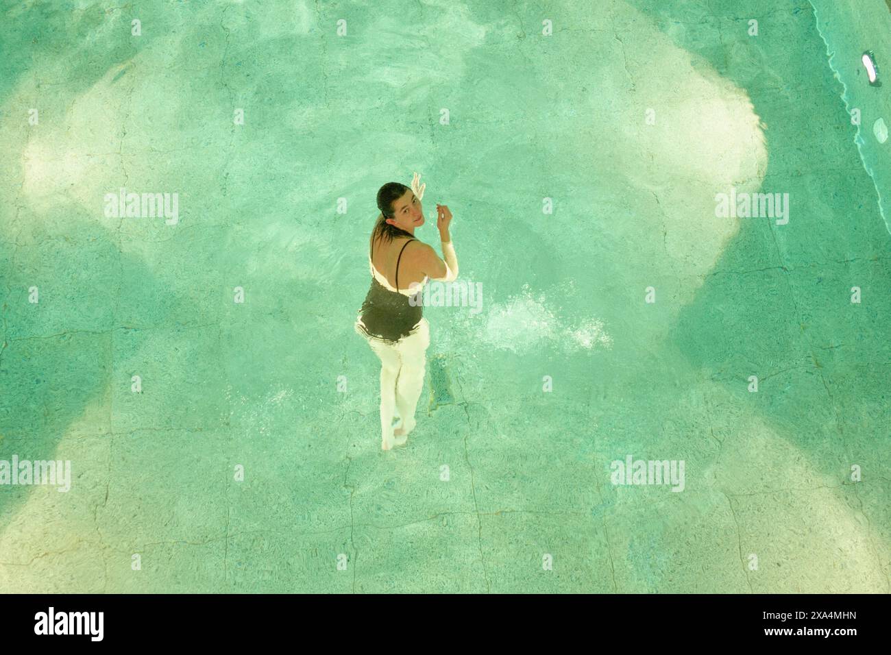 Eine Frau steht in einem Pool mit klarem blauem Wasser und blickt mit einer Hand leicht angehoben zur Kamera. Stockfoto
