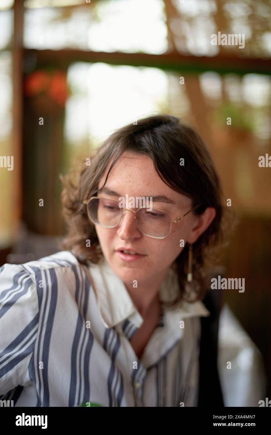 Ein junger Erwachsener mit Brille und kurzen Haaren schaut nachdenklich nach unten, trägt ein gestreiftes Hemd und Overall vor einem verschwommenen Hintergrund. Stockfoto